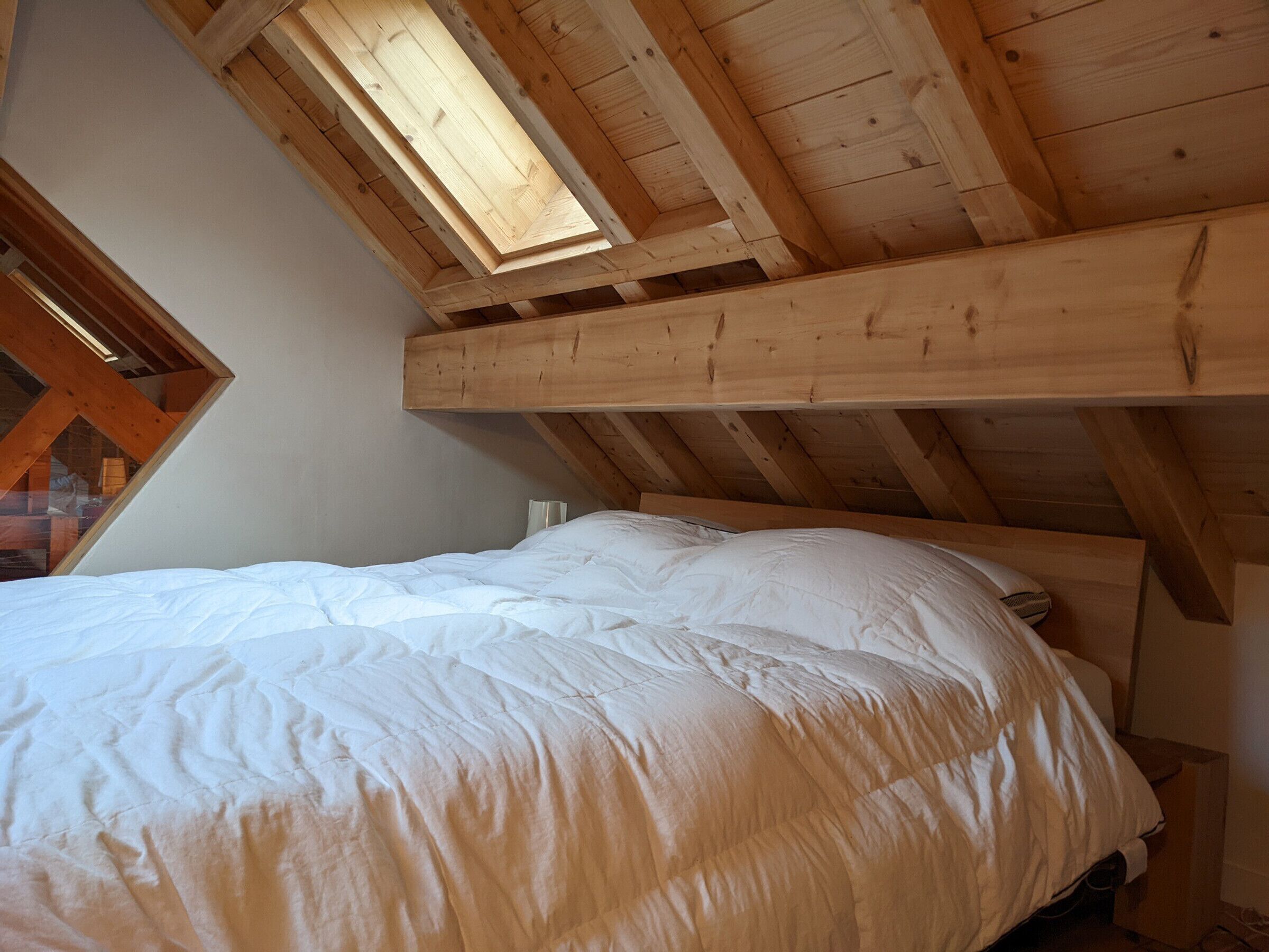 A bedroom with a sloped ceiling, wooden beams, a bed with white bedding, and a skylight.