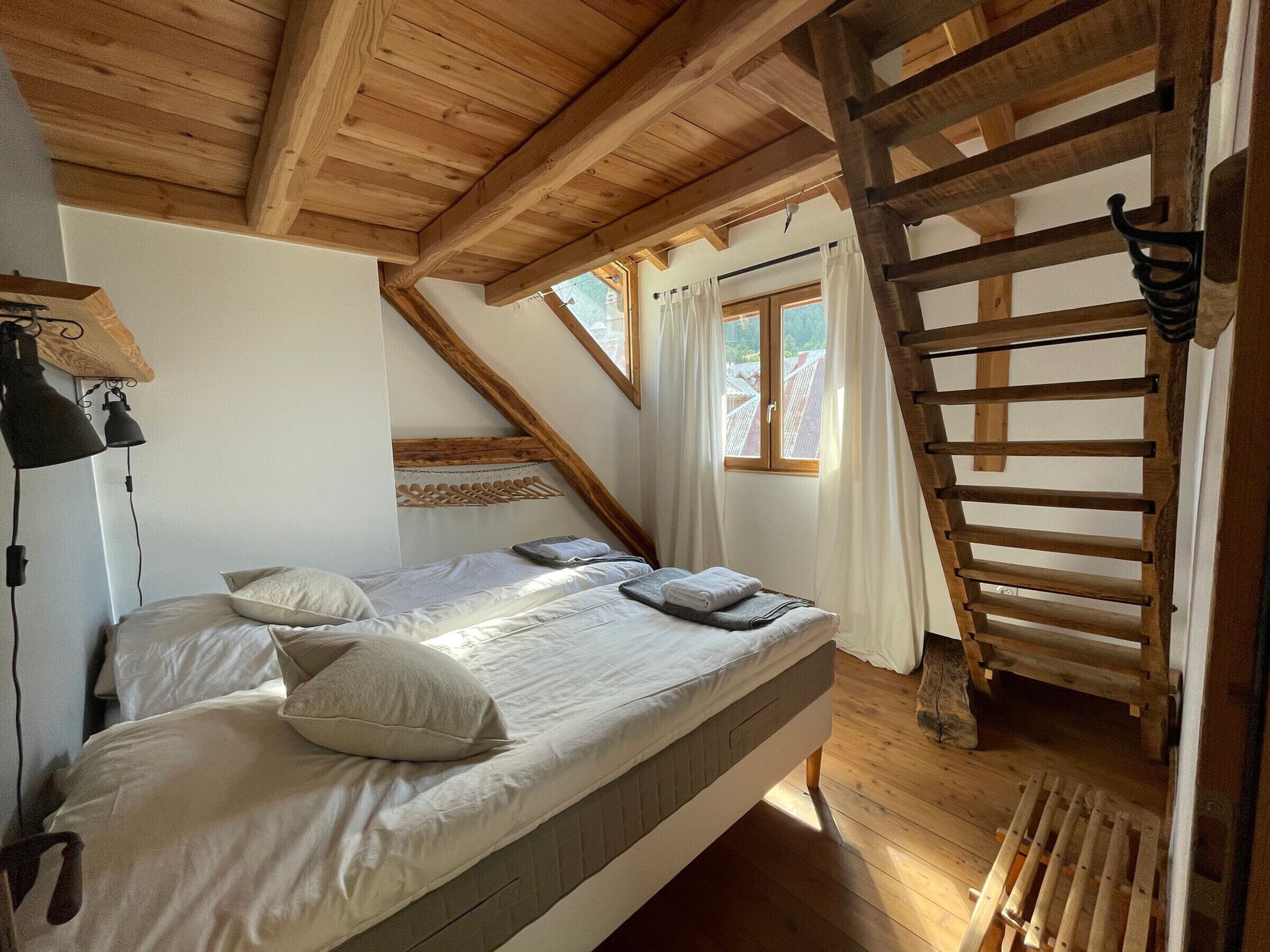 Een slaapkamer met een hoogslaper, houten balken, een raam met gordijnen en een houten trap.