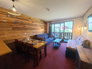 A cozy room with a wooden wall, a dining table with chairs, a sofa, and a small table with a lamp.