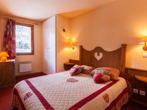 A bed with a quilt, a nightstand, a wall-mounted lamp, and a window with curtains.