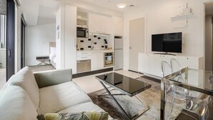 Ein modernes Wohnzimmer mit einer Couch, einem Couchtisch und klaren Glaskästen. Im Hintergrund ist eine Küche mit weißen Schränken und einer Mikrowelle zu sehen.