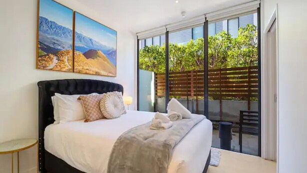 A bedroom with a large bed, two wall-mounted paintings, a sliding glass door leading to an outdoor area with greenery. - Charming 1BR in Wynyard Quarter I Zodiak Stays (Auckland)