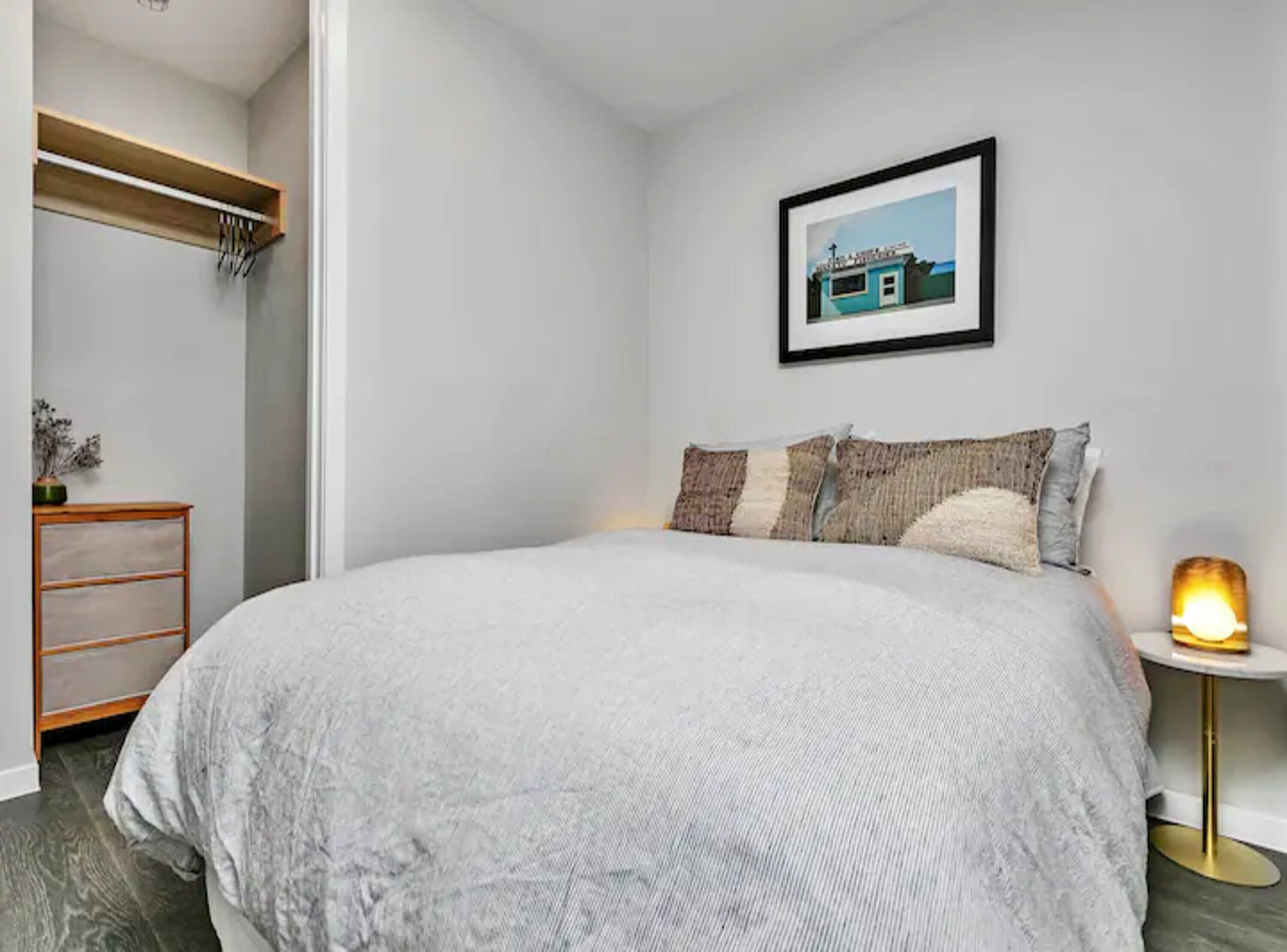 A bedroom with a large bed, a bedside table with a lamp, a framed picture on the wall, and a wooden shelf with hanging items.