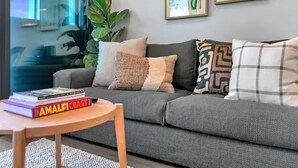 Un salon moderne avec un canapé gris, une petite table en bois sur laquelle sont posés des livres, et deux œuvres d’art encadrées accrochées au mur.
