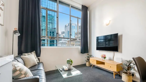 A modern living room with a leather sofa, a glass coffee table, a wooden TV stand, and a large window offering a city view.