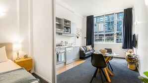 A modern hotel room with a bed, a desk, a dining area, a kitchenette, and a large window with curtains.