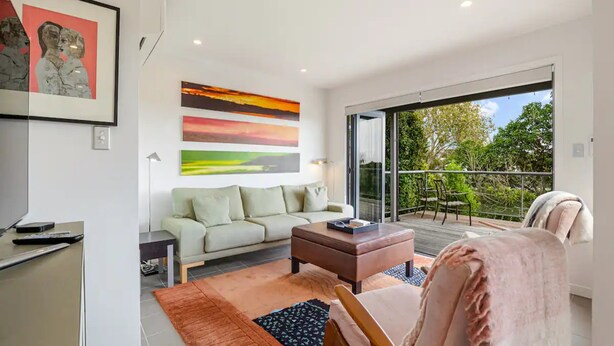 A modern living room with a sofa, a coffee table, and a chair. There is a painting on the wall and a balcony with a view of trees. - Stylish And Serene Living In Kohimarama (Auckland)