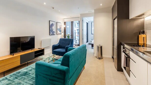 A modern apartment with a living area featuring a sofa, a TV, and a glass coffee table. There is a kitchen with a stove and a trash can. - Central & Modern 2-Bedroom Apartment (Auckland)