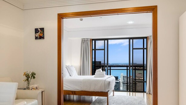 Une chambre d’hôtel avec un lit, un canapé blanc, une vue sur la ville grâce à de grandes fenêtres et un balcon donnant sur la mer.