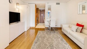 Un salon moderne doté d’un téléviseur à écran plat, d’un canapé blanc agrémenté de coussins à motifs, d’un tapis à motifs et d’un coin repas avec une table et des chaises blanches.