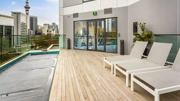 Un edificio moderno con zona de piscina en la azotea, sillones de descanso y entrada por puerta de cristal.