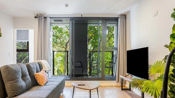 A modern living room with a grey sofa, a coffee table, a TV, and a balcony with a view of trees. - Auckland City Charm - 2BR Haven by Zodiak Stays (Auckland)