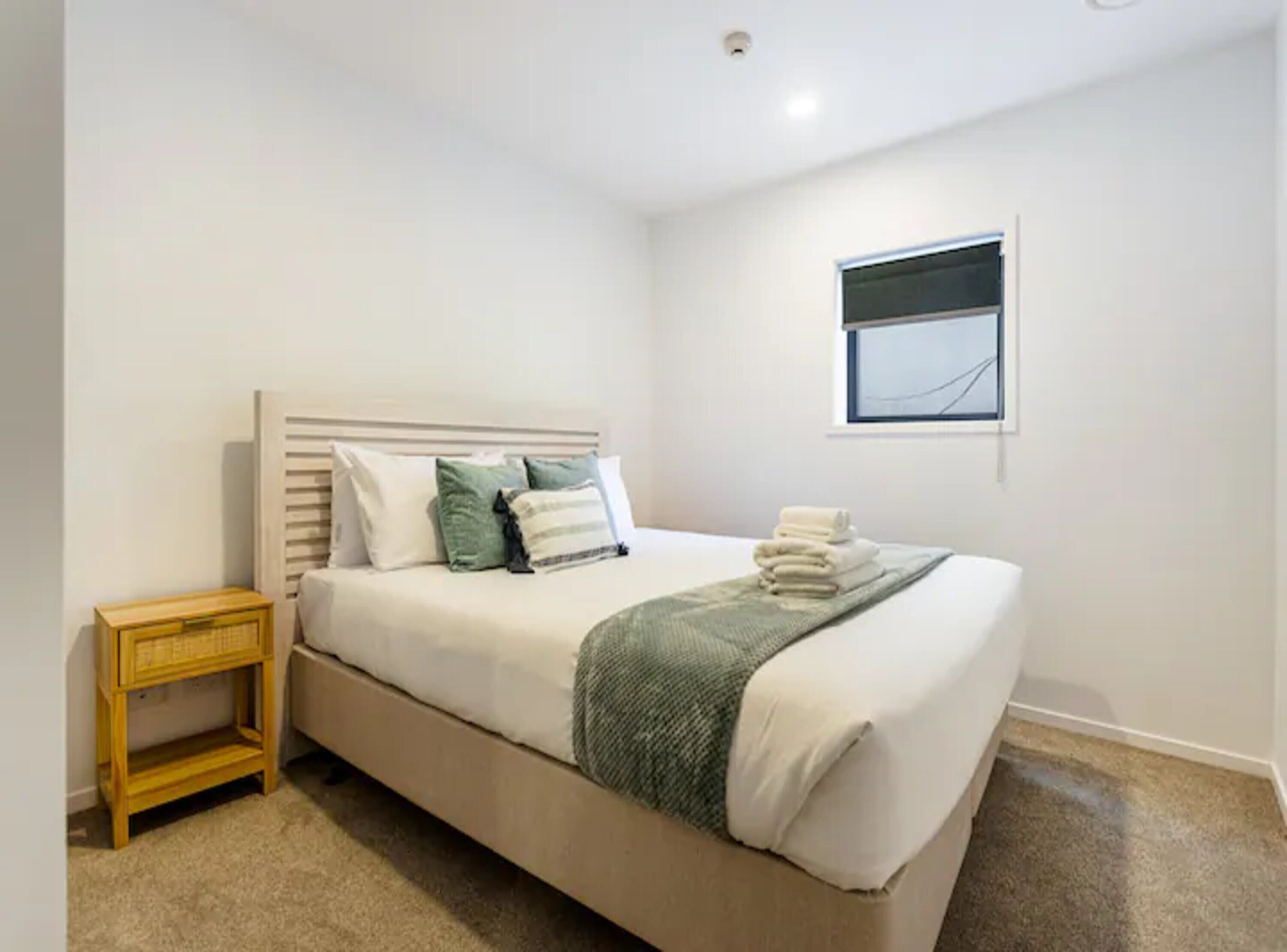Una cama bien hecha con almohadas y una colcha, una mesita de noche de madera y una ventana con persianas.