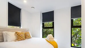 A hotel room with a large bed, two windows with black blinds, and a view of greenery outside. - Auckland City Charm - 2BR Haven by Zodiak Stays (Auckland)