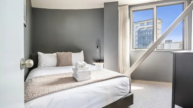Een moderne hotelkamer met een groot bed, een nachtkastje, een lamp en uitzicht op de stad door een groot raam.