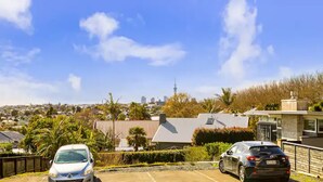 A residential area with two cars parked in a driveway, a clear blue sky, and a distant view of a city skyline including a tall tower.