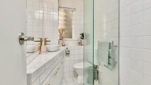 A modern bathroom with white tiles, a glass shower enclosure, and a vanity with a sink and mirrors. - Heart Of The City Apartment In Heritage Building (Auckland)