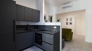Eine moderne Küche mit dunklen Schränken, einem schwarzen Spülbecken und einem eingebauten Backofen. Im angrenzenden Wohnbereich befindet sich ein grünes Sofa, und der Raum ist in einer neutralen Farbgebung gehalten.