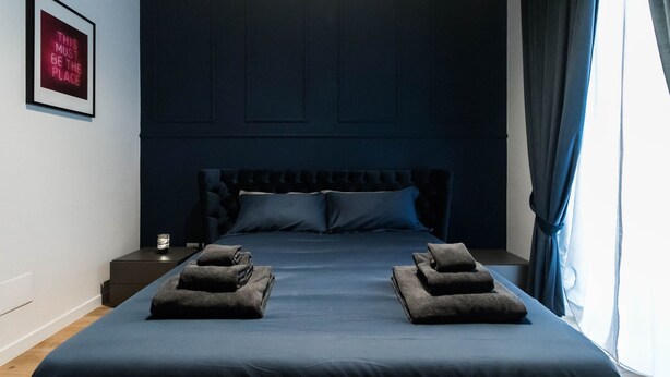 Ein ordentlich bezogenes Bett mit dunklem Kopfteil und Kissen, ein Nachttisch mit Lampe sowie ein gerahmtes Schild an der Wand.