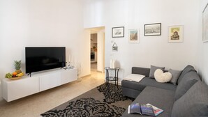 Een moderne woonkamer met een flatscreen televisie op een witte kast, een grijze bank met een hartvormig kussen en een zwart-wit tapijt met een patroon.