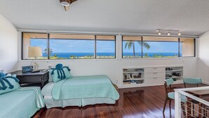 A bedroom with a large window, wooden flooring, a bed with teal bedding, a desk, and a chair. - 🌴 Newly Renovated Top Floor Condo with Golf Course & Ocean Views 🌴 (Kailua Kona)