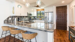 A modern kitchen with a large island, bar stools, hanging pendant lights, and a view of the outdoors. - 🌴 Newly Renovated Top Floor Condo with Golf Course & Ocean Views 🌴 (Kailua Kona)