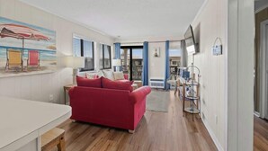 A spacious living room with a red sofa, a dining area, and a view of the city through large windows. - Ocean Forest Villas Unit C301 - Ocean view (Myrtle Beach)