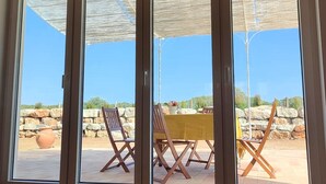 Een kamer met een schuifglasdeur die uitkomt op een terras met een gele tafel en stoelen, een grote terracotta pot en een stenen muur.