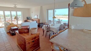 Een ruime woonkamer met een groot raam met uitzicht op een strand, een eethoek met tafel en stoelen, en een gezellige zithoek met een bank en een open haard.