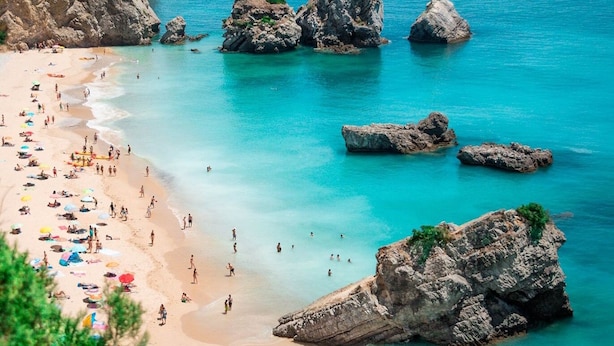 Een strand met helder turquoise water, rotsformaties en parasols.