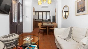 Ein Wohnzimmer mit Sofa, einem Glastisch, einem Fernseher, einer Essecke mit Tisch und Stühlen sowie einer Küche im Hintergrund.