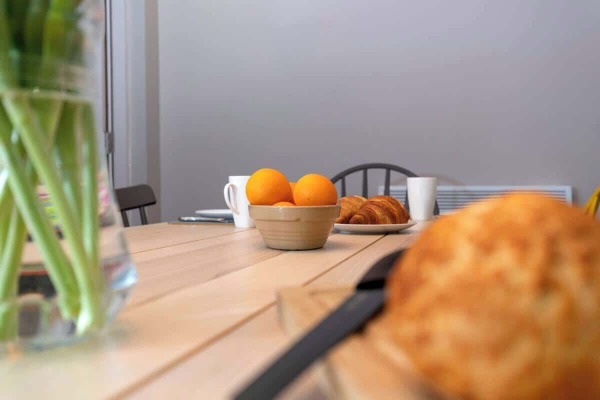 Een eettafel met een schaal oranjes, een croissant, een witte mok en een stoel.