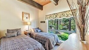 A bedroom with two beds, a bedside table with a lamp, a window with a view of greenery, and a wooden table with a vase. - 45 Coeur Du Lac 3BR/2BA Incline Village, Pool & Mtn View (Incline Village)