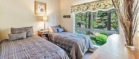 A bedroom with two beds, a bedside table with a lamp, a window with a view of greenery, and a wooden table with a vase.