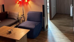 Un salon moderne avec un canapé, un fauteuil bleu, une table basse en bois et un lampadaire avec des lumières rouges. Un couloir mène à une chambre dotée d’une armoire et d’un ventilateur de plafond.