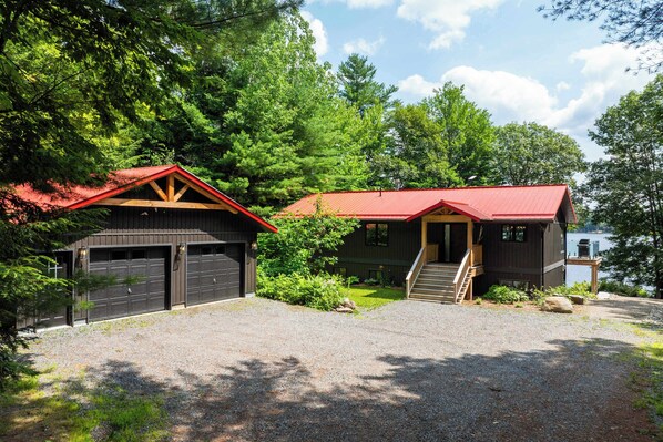 A two-story cabin with a red roof, a gravel driveway, and a lake view.
