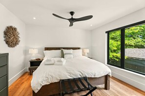 A bedroom with a bed, bedside tables, a ceiling fan, a window with a view of greenery, and a wall-mounted artwork.