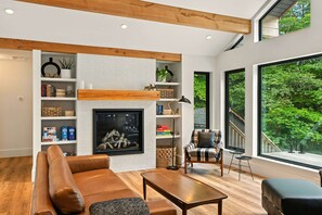 Een moderne woonkamer met een open haard, houten balken, een groot raam met uitzicht op bomen en een gezellige zithoek.
