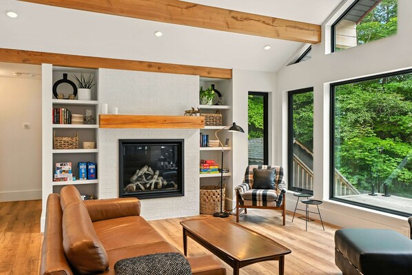 A modern living room with a fireplace, wooden beams, a large window with a view of trees, and a cozy seating area. - Making Memories (Baysville)