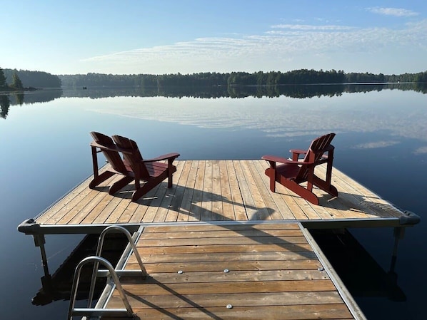 A wooden dock with two red Adirondack chairs facing a calm lake.