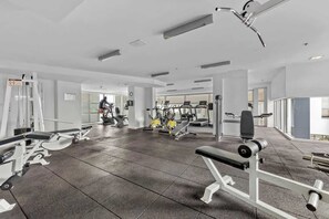Une salle de sport moderne équipée de divers appareils d’exercice, une personne qui s’entraîne et de grandes fenêtres laissant entrer la lumière naturelle.