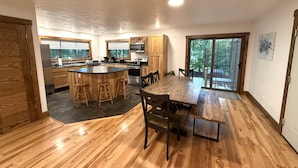 A modern kitchen with wooden cabinets, a central island with stools, a dining area with a large table and chairs, and a view through a glass door. - Wooded Bliss @ Spring Brook Resort (Wisconsin Dells)