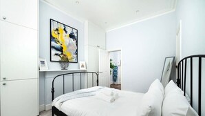 Un dormitorio con cama, paredes blancas, un cuadro y una puerta que da a otra habitación.