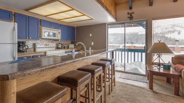 Cocina moderna con armarios azules, una isla central con taburetes, microondas y un amplio ventanal con vistas a un paisaje nevado.