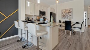 Uma cozinha moderna com armários brancos, uma ilha central com banquetas, uma área de refeições com mesa e cadeiras, e um grande quadro abstrato na parede.