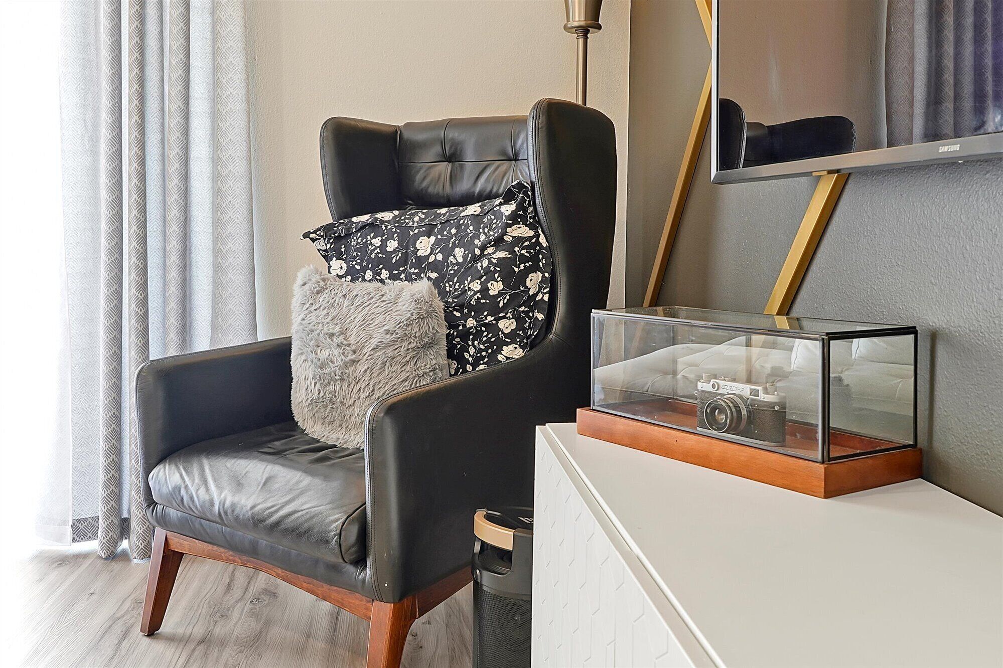 Een moderne woonkamer met een zwarte leren fauteuil, een zacht grijs kussen en een vintage camera in een glazen displaykast.
