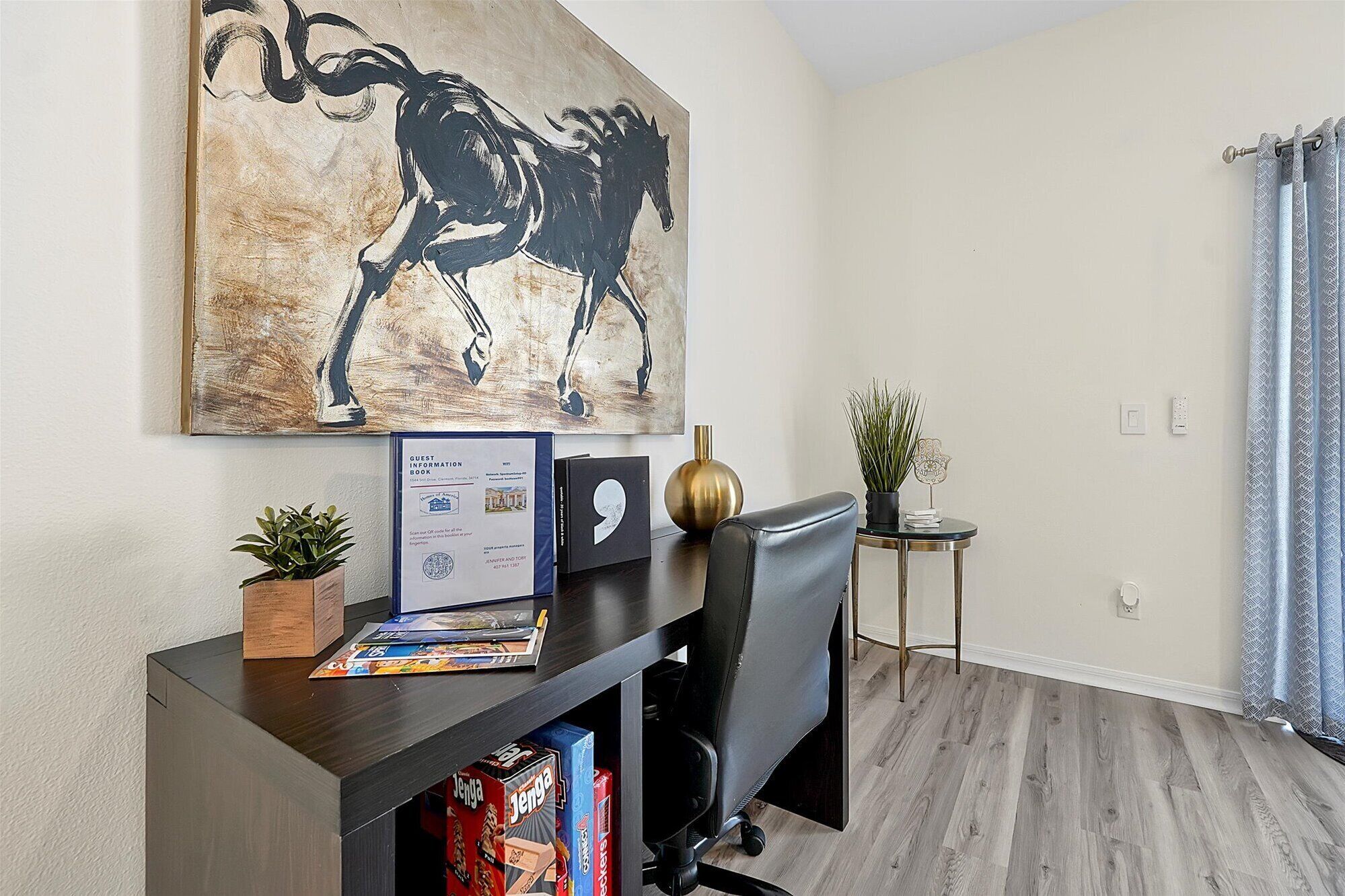A modern office space with a large abstract painting, a desk with a chair, a small side table with a plant, and a bookshelf.