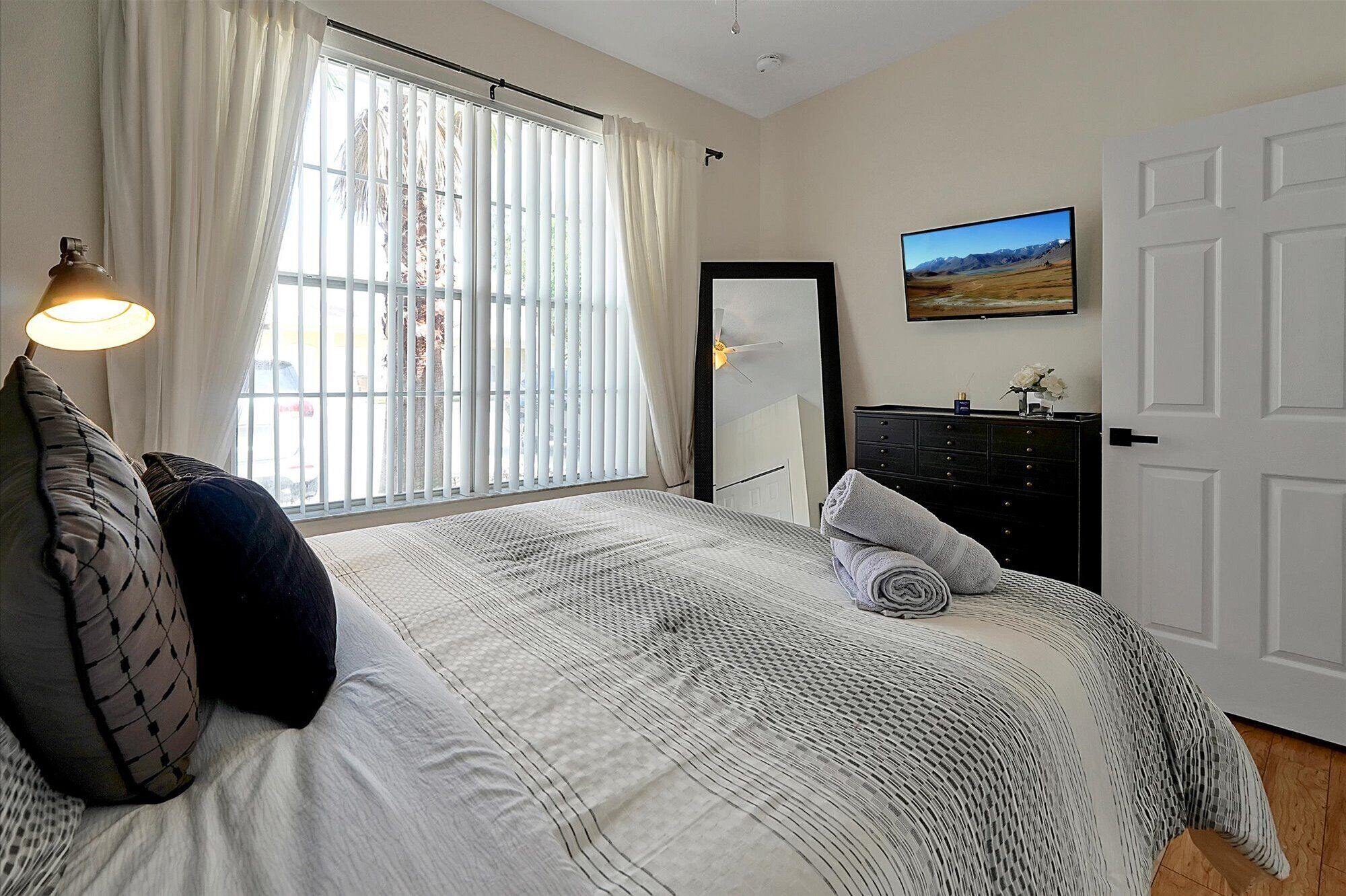 Une chambre à coucher avec un lit, une commode, un miroir et une fenêtre avec des rideaux.