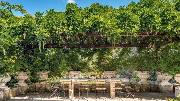 Un espacio al aire libre con mobiliario, una pérgola cubierta de vegetación, macetas de piedra y una mesa de madera rústica con sillas.