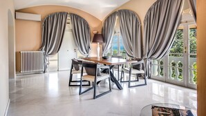 Un comedor con mesa y sillas, ventanas amplias con cortinas, un radiador y vistas a zonas verdes.